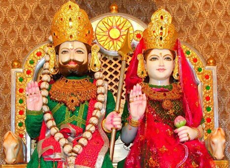 Pair of Hindu deity statues on an ornate throne, adorned with gold crowns, jewelry, and bright robes, raising one hand each in blessing.