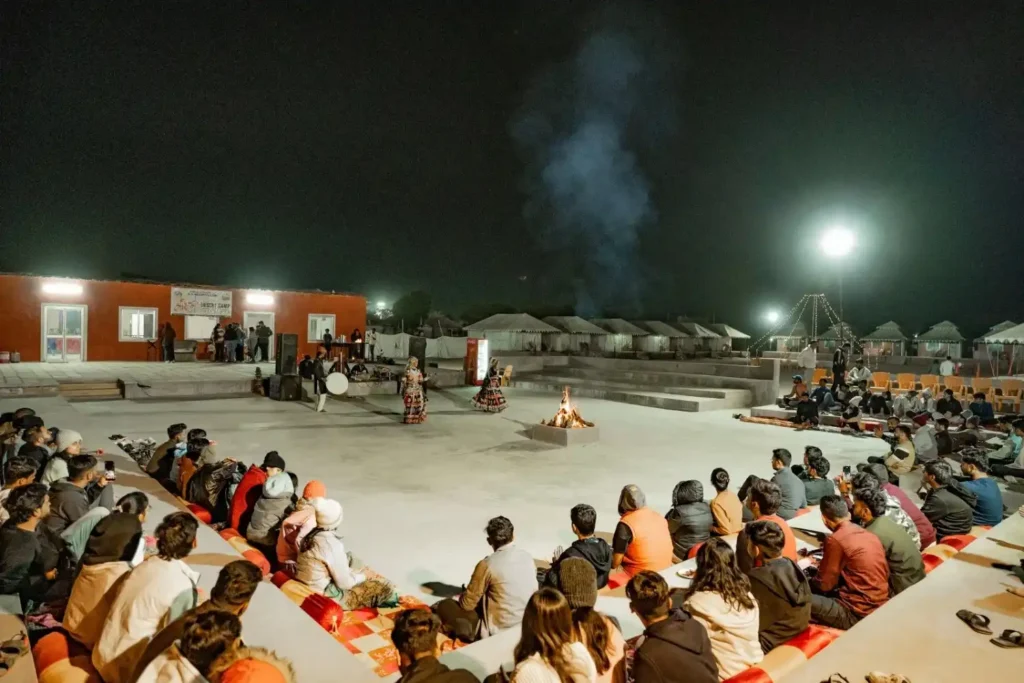 Nighttime outdoor festival with a central bonfire, dancers performing in front of an audience seated on steps and mats around the fire pit.