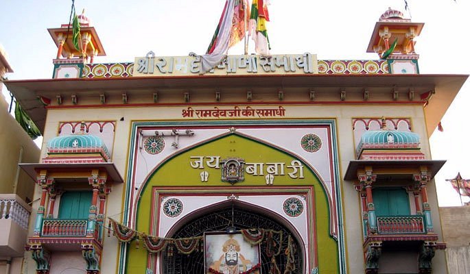 Colorful Hindu temple façade with ornate arches, balconies, and Hindi lettering above the entrance, a framed portrait displayed at the doorway.