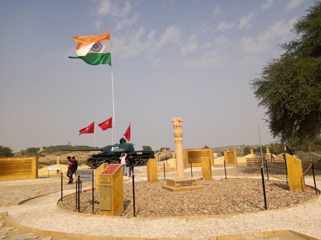 Indian flag atop a military tank displayed at a war memorial with surrounding plaques and signage in a sunny open area.