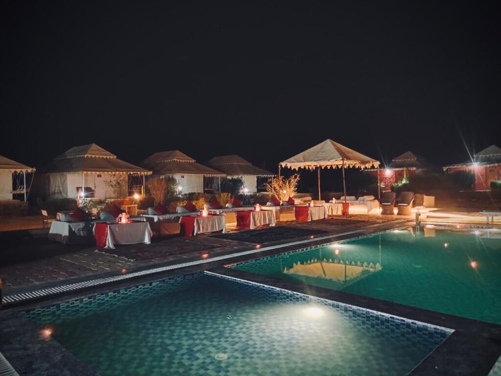 luxury camp in jaisalmer - Shrinath Desert Camp Jaisalmer Nighttime poolside event with white tents, lanterns, and tables dressed in red and white along the pool edge