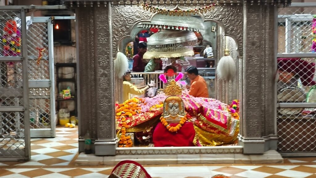 Ornate temple shrine with a garlanded deity statue surrounded by colorful fabrics, worshippers nearby