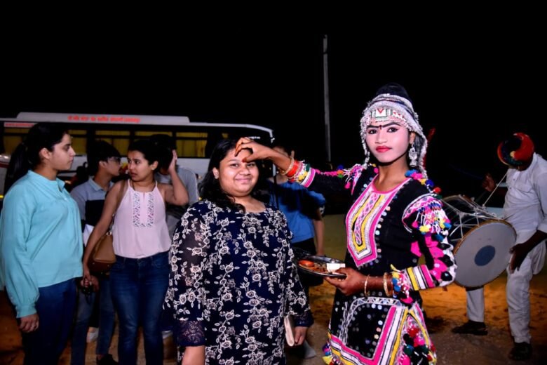 A woman in a colorful traditional dress applies a red tilak on another woman's forehead at night, with bystanders and a drummer in the background.