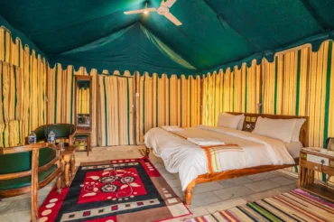 Glamping tent interior with a wooden king bed, white bedding, striped yellow-green walls, and a ceiling fan above.