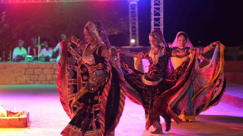 Women in vibrant traditional Indian outfits perform a choreographed dance with flowing dupattas on an outdoor stage at night.